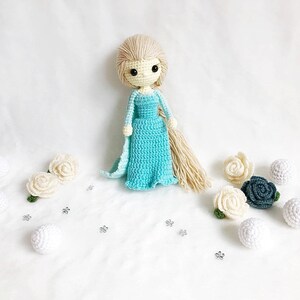 May include: A crocheted doll with long blonde hair wearing a light blue dress and holding a white crocheted flower. The doll is surrounded by white and green crocheted flowers.