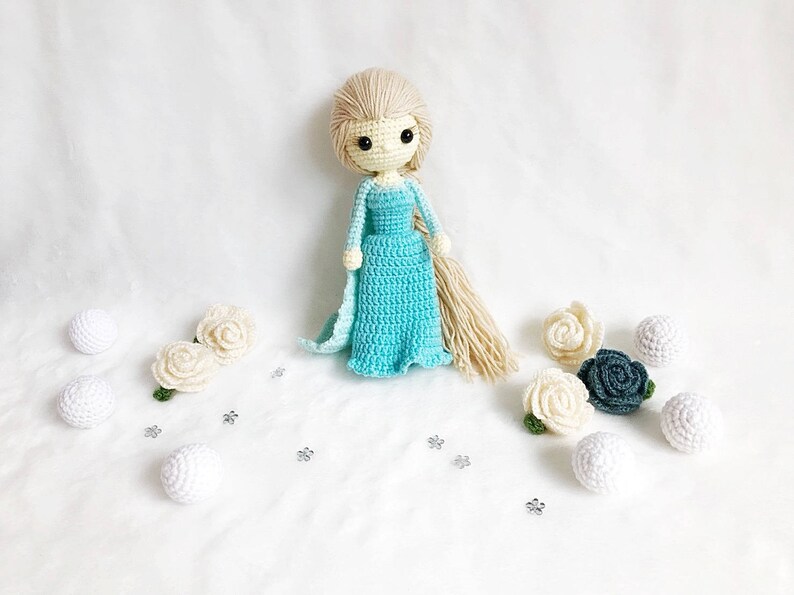 May include: A crocheted doll with long blonde hair wearing a light blue dress and holding a white crocheted flower. The doll is surrounded by white and green crocheted flowers.