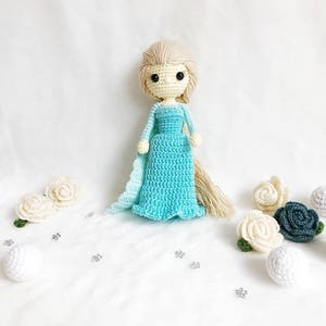 May include: A crocheted doll with blonde hair wearing a light blue dress and a white cape. The doll is surrounded by white and blue crocheted flowers and white balls.