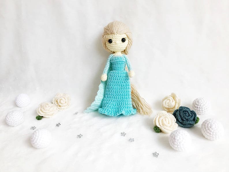 May include: A crocheted doll with blonde hair wearing a light blue dress and a white cape. The doll is surrounded by white and blue crocheted flowers and white balls.