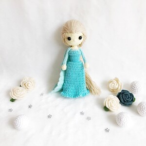 May include: A crocheted doll with long blonde hair wearing a light blue dress and standing on a white surface with white and blue crocheted flowers.