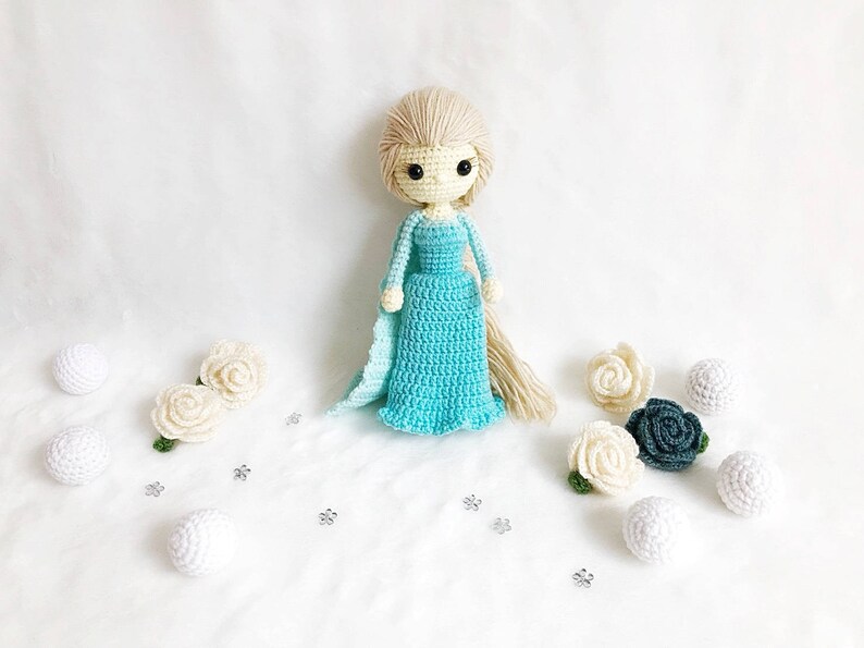 May include: A crocheted doll with long blonde hair wearing a light blue dress and standing on a white surface with white and blue crocheted flowers.