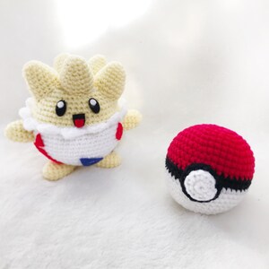 May include: Handmade crochet Togepi and Pok&eacute; Ball. The Togepi is cream, white, red, and blue. The Pok&eacute; Ball is red, black, and white. The Togepi has a happy expression. The items are on a white surface.