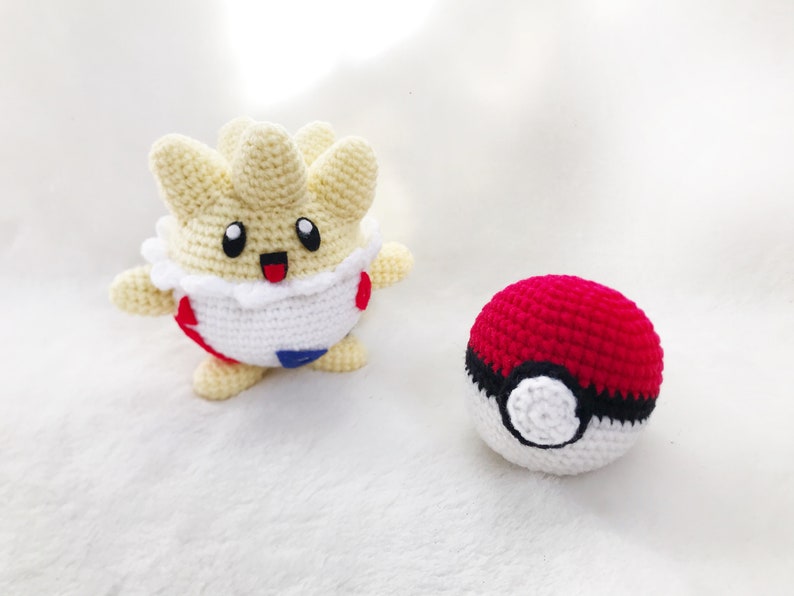 May include: Handmade crochet Togepi and Pok&eacute; Ball. The Togepi is cream, white, red, and blue. The Pok&eacute; Ball is red, black, and white. The Togepi has a happy expression. The items are on a white surface.