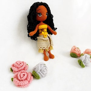 May include: Crocheted doll with brown skin, black curly hair, and a yellow and brown top. The doll is wearing a beige skirt and is surrounded by crocheted flowers in pink, gray, and green.