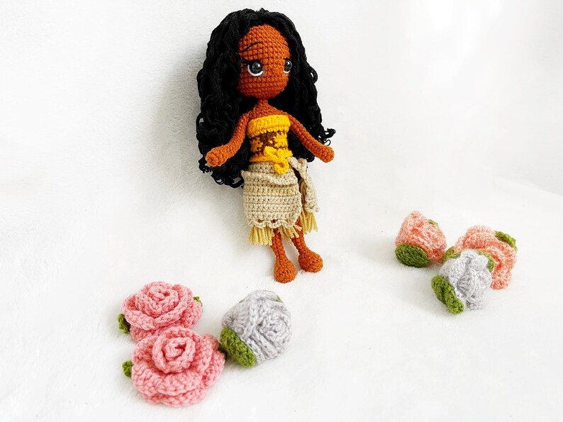 May include: Crocheted doll with brown skin, black curly hair, and a yellow and brown top. The doll is wearing a beige skirt and is surrounded by crocheted flowers in pink, gray, and green.