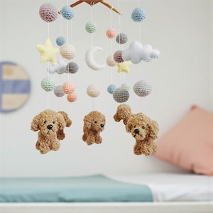 May include: A handcrafted baby mobile with crocheted components. The mobile showcases brown dog figures, colourful spheres, stars, clouds, and a crescent moon. It hangs from a wooden frame, offering a whimsical nursery decoration.