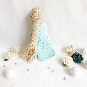 May include: A crocheted doll with long blonde hair in a braid, wearing a light blue dress. The doll is surrounded by white and green crocheted flowers and white balls.