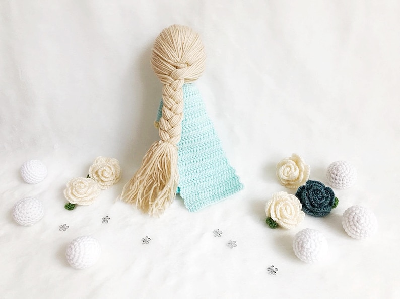 May include: A crocheted doll with long blonde hair in a braid, wearing a light blue dress. The doll is surrounded by white and green crocheted flowers and white balls.