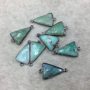 May include: A collection of triangular gemstone pendants in shades of green and blue. Each pendant has a silver-colored metal frame and small loops at the top and bottom for attaching to jewelry. The gemstones have a faceted appearance.