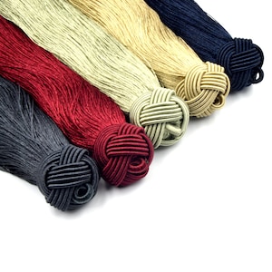 May include: Five tassels in different colors, including red, green, beige, gold, and navy blue, with knotted ends. The tassels are made of silk and are arranged in a row on a white background.