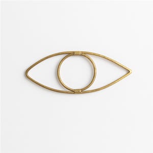 Könnte beinhalten: Goldfarbene Wanddekoration in Form eines Auges aus Metall. Das Design zeigt eine äußere Augenform mit einer kreisförmigen Iris in der Mitte. Das Metall hat eine leicht strukturierte Oberfläche vor weißem Hintergrund.