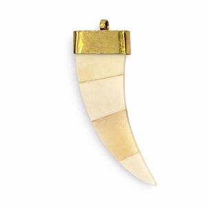 Puede incluir: Colgante de color marfil en forma de diente curvado, con una tapa y un lazo de metal dorado. El colgante está segmentado con tonos marfil más claros y oscuros, creando un efecto rayado. El diseño es simple y elegante.