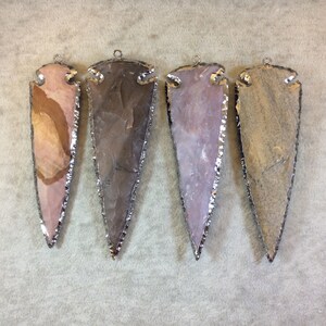 May include: Four arrowhead pendants with silver-toned metal trim. The arrowheads are made of natural stone with variations in color and pattern. The arrowheads are approximately 2 inches in length.