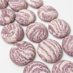 May include: A collection of round, smooth objects featuring a marbled pattern of creamy white and speckled purple. Each piece has a distinct, swirling texture with varying concentrations of purple pigment across the surface, creating a cohesive set.