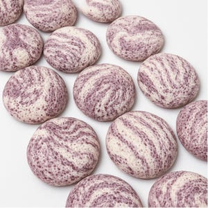 May include: A collection of round, flat stones with a marbled appearance. The stones feature a cream-colored base with swirling patterns of deep purple. The arrangement is on a white background, highlighting the natural textures and colors of the stones.