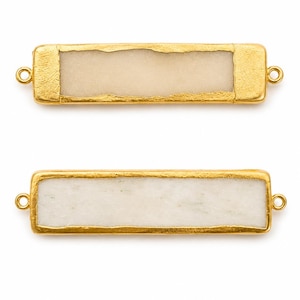 Puede incluir: Dos conectores rectangulares para joyería con un marco de metal dorado texturizado y una incrustación central de piedra lisa en color blanco hueso. Cada pieza tiene un pequeño aro circular en ambos extremos para sujetar cadenas.