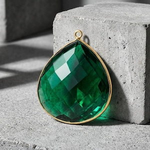 May include: A large, faceted emerald-green gemstone pendant with a gold-colored bezel. The teardrop-shaped jewel has a small loop at the top for hanging. The pendant is displayed on a textured gray surface.