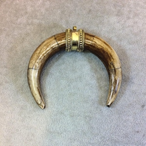 May include: A curved pendant made of bone-colored material with brown accents. The pendant has a gold-colored metal band with a decorative design at the top. The pendant is a crescent shape.