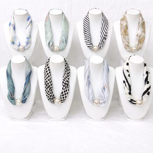 May include: Eight silk scarves in various patterns and colors, displayed on white mannequin busts. The scarves feature designs such as floral, polka dots, stripes, and abstract prints. Each scarf is secured with a large, round, pearl-like clasp.
