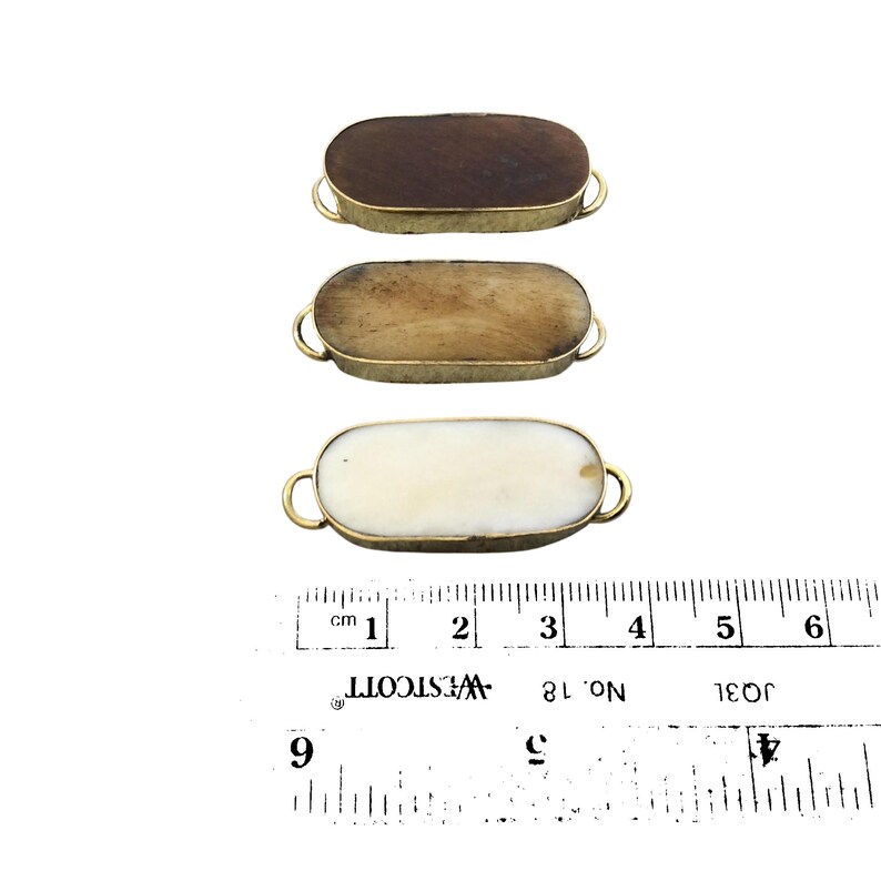 Może przedstawiać: Trzy owalne guziki z kości i mosiądzu. Guziki mają około 5 cm długości i 2,5 cm szerokości. Kość jest w naturalnym kolorze, z niewielkimi r&oacute;żnicami w kolorze i fakturze. Mosiądz jest błyszczący, w kolorze złotym.