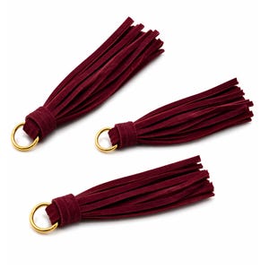 Puede incluir: Tres borlas de ante burdeos con anillos de metal color oro. Las borlas están dispuestas sobre un fondo blanco. Cada borla tiene un lazo para sujetarla a un llavero, cremallera u otro proyecto de manualidades. Las borlas son de color rojo oscuro.