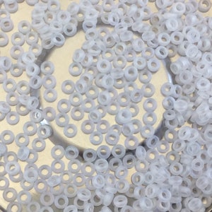 May include: A close-up image of a large quantity of small, translucent white glass beads. The beads are shaped like rings and are scattered on a light gray surface.
