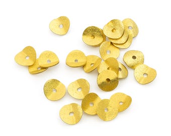 May include: A collection of small, circular gold-colored metal sequins or beads, each featuring a single central hole. The pieces have a slightly irregular, wavy edge and a textured, matte finish, scattered across the frame in a loose arrangement.