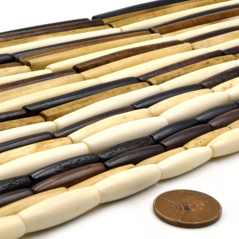 Pu&ograve; includere: Primo piano di una collana di perline in osso in varie tonalit&agrave; di marrone, beige e bianco. Le perline sono di forma ovale e hanno una finitura liscia e lucida. Un penny &egrave; incluso per la scala.