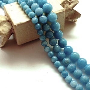 Peut inclure: Plusieurs brins de perles de pierres précieuses d'aigue-marine bleu clair de différentes tailles. Les perles rondes ont une finition polie et brillante. Les perles sont enfilées, prêtes pour la fabrication de bijoux. Une petite boîte en carton marron est en arrière-plan.