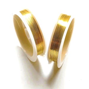 Op de afbeelding: Twee spoelen goudkleurige draad worden getoond tegen een witte achtergrond. De draad is strak opgerold op witte plastic spoelen. De draad heeft een glanzende, metallic uitstraling, wat suggereert dat het van een edelmetaal of een goudkleurige legering is gemaakt. Ideaal voor het maken van sieraden.