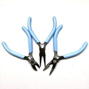 May include: Four metal pliers with black jaws and light blue handles are arranged on a white surface. The pliers include needle-nose, round-nose, and side-cutting styles, commonly used for jewellery making and crafting.