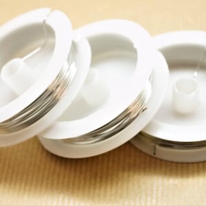 Peut inclure: Trois bobines de fil argenté sont présentées, chacune enroulée autour d'une bobine en plastique blanc. Les bobines sont disposées en rangée, le fil étant visible sur les bords. L'arrière-plan est de couleur beige clair.