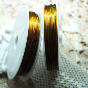 May include: Two spools of gold-coloured beading wire. The wire is tightly wound on white plastic spools. The spools are positioned close together, with the wire strands visible. The background is a soft, textured surface.