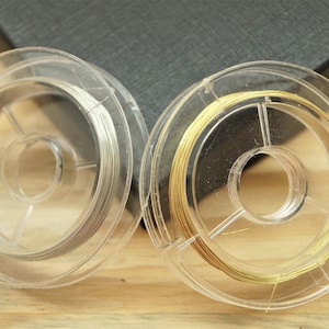 Può includere: Sono mostrate due bobine di filo per gioielli. Una bobina contiene filo color argento, l'altra filo color oro. Entrambe le bobine sono di plastica trasparente e hanno una forma circolare. Lo sfondo è di colore grigio scuro.