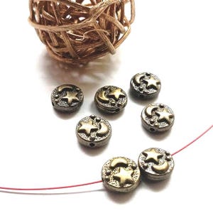 Peut inclure: Perles rondes de couleur bronze antique avec des motifs d'étoiles et de croissants de lune. Les perles sont dispersées sur une surface blanche, avec une boule tissée en arrière-plan. Idéales pour la création de bijoux et l'artisanat.