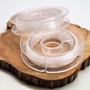 May include: Two spools of clear elastic string wound around plastic spools. The spools are stacked on top of each other and are sitting on a wooden surface.