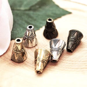 Puede incluir: Un surtido de cuentas cónicas decorativas de metal en varios acabados metálicos, incluyendo oro, plata y bronce. Las cuentas presentan intrincados patrones en relieve y están diseñadas para la fabricación de joyas o proyectos de artesanía. Las cuentas se muestran sobre una superficie de madera clara.