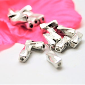 Könnte beinhalten: Eine Gruppe von silbernen Metallperlen mit einem gedrehten Design. Die Perlen haben jeweils zwei Löcher und sind auf einer weißen Oberfläche verstreut.