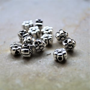 Peut inclure: Un ensemble de petites perles en métal argenté en forme de fleurs à cinq pétales. Chaque perle présente un trou central pour l'enfilage et une finition métallique texturée avec des zones sombres en creux pour souligner les détails des pétales.