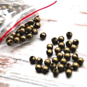 Può includere: Una collezione di piccole perline rotonde color ottone antico. Alcune perline sono in un sacchetto di plastica trasparente con una cerniera rossa, mentre altre sono sparse su una superficie bianca. Le perline hanno un foro al centro.