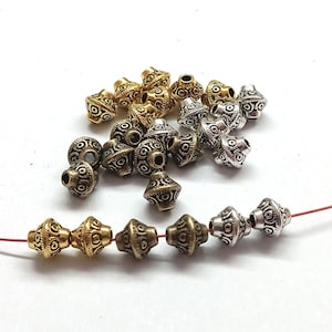Puede incluir: Una colección de cuentas decorativas de metal en tonos dorados, plateados y bronce. Las cuentas tienen un diseño único y estampado y están ensartadas en un cordón rojo. Ideales para la fabricación de joyas y proyectos de artesanía.