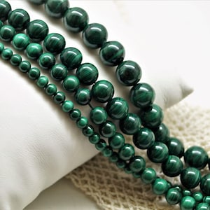 Peut inclure: Quatre rangées de perles de malachite vert foncé de tailles variées sont présentées. Les perles sont rondes et polies, avec un motif tourbillonnant. Les rangées sont disposées en diagonale sur un fond blanc.