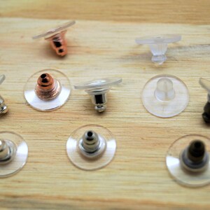 Peut inclure: Gros plan de divers fermoirs de boucles d'oreilles, comprenant des disques en plastique transparent et des tiges métalliques argentées, cuivrées et noires. Les fermoirs sont disposés sur une surface en bois brun clair. Ces apprêts sont pour la création de bijoux.