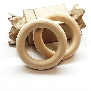 May include: Two light brown wooden rings are shown on a white surface. The rings are circular with a thick, rounded profile. A small cardboard box and a piece of brown paper are in the background. The rings are likely for crafts.