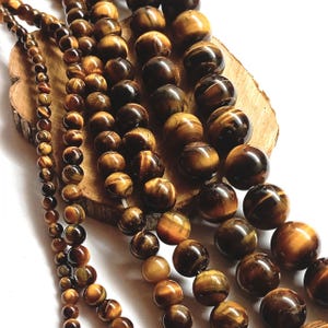 Peut inclure: Plusieurs rangées de perles en œil-de-tigre polies de tailles variées, allant de petites à grandes sphères. Les perles présentent un éclat chatoyant caractéristique avec des bandes alternées de couleurs brun doré et brun foncé, disposées en lignes.