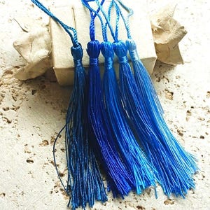 Puede incluir: Un conjunto de seis borlas azules de diferentes tonos, desde azul marino oscuro hasta azul celeste brillante. Cada borla está hecha de hilos sedosos, con una parte superior en bucle para colgar. Las borlas están dispuestas sobre una superficie clara y texturizada.