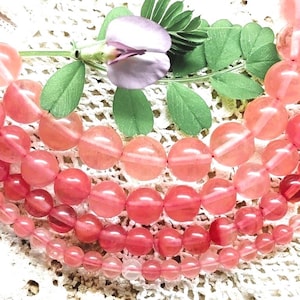 Peut inclure: Gros plan sur plusieurs rangées de perles de pierres précieuses roses et rouges. Les perles sont rondes et translucides. Une branche de feuilles vertes et une fleur violette sont dans la partie supérieure de l'image.