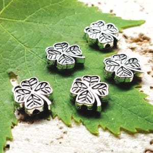 May include: Five silver clover charms are shown on a green leaf. Each charm features four detailed leaves with visible veins. The charms are scattered across the leaf, creating a visually appealing arrangement. The background is a mix of green and beige.