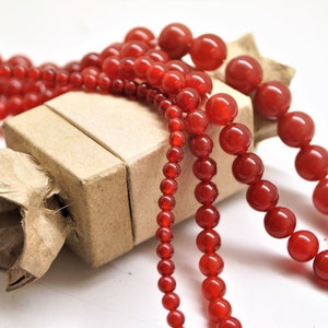 Puede incluir: Primer plano de tres hebras de cuentas de cornalina roja. Las cuentas son redondas y lisas, y están dispuestas en un montón suelto sobre una caja de cartón marrón.